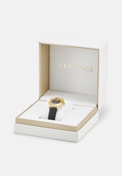 Black watch with gold accents, round face, Greek key pattern around bezel, presented in a white and gold box with a soft interior.