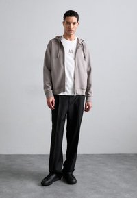 Grå sweatshirt med hætte og lynlås foran, parret med en hvid T-shirt med et lille logo, og sorte bukser. Sorte sneakers fuldender looket.