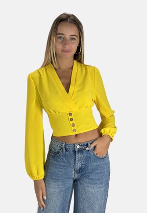 Gelbe Wickelbluse mit langen Puffärmeln, die eine taillierte Taille und drei dekorative Knöpfe aufweist. Kombiniert mit hochtaillierten blauen Jeans.