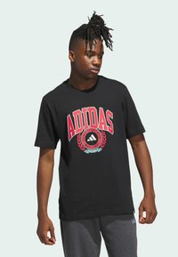 Černé bavlněné tričko s velkým červeným logem Adidas, kulatý znak se designem vavřínového věnce, krátké rukávy. Nosí se s šedými kalhotami.