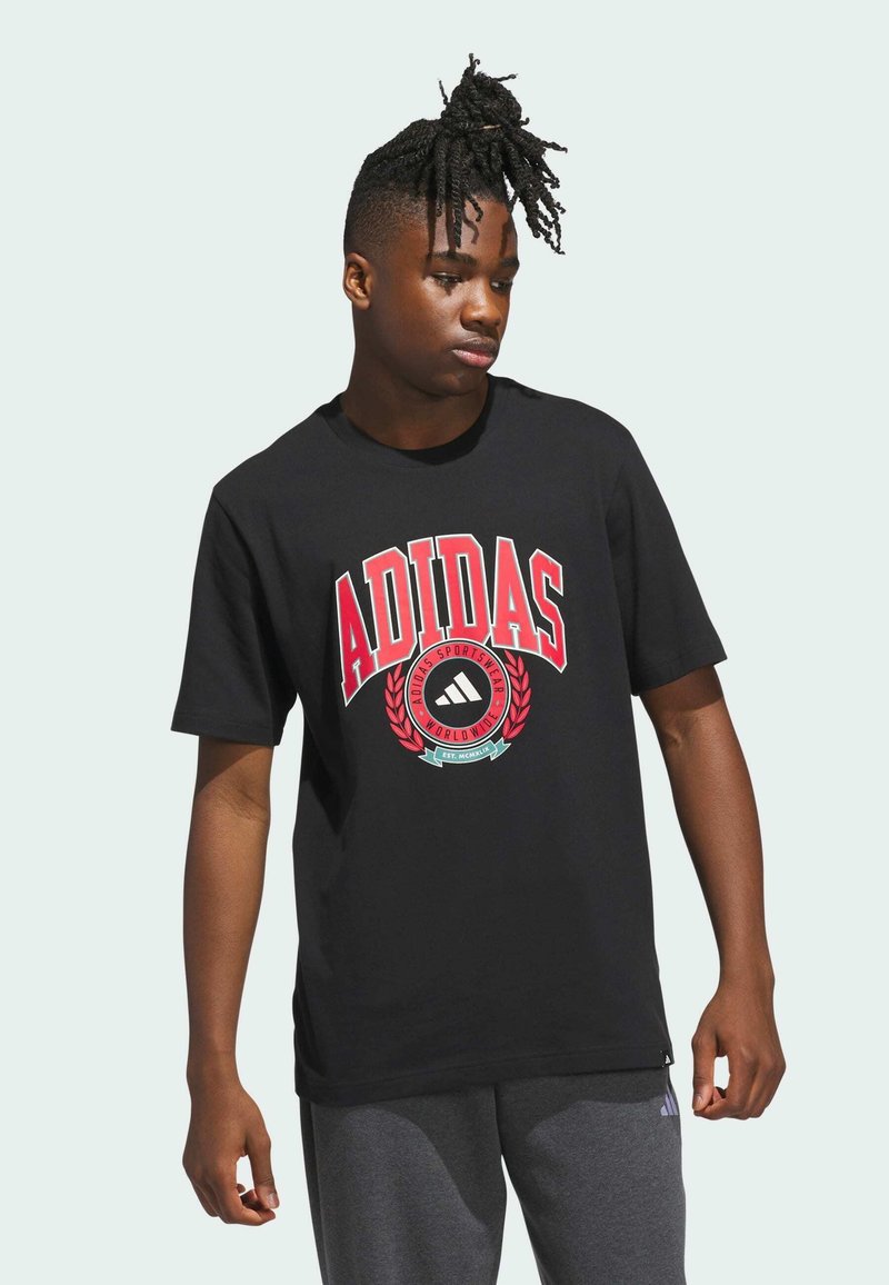 Černé bavlněné tričko s velkým červeným logem Adidas, kulatý znak se designem vavřínového věnce, krátké rukávy. Nosí se s šedými kalhotami.