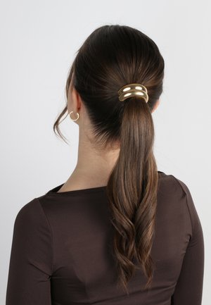Fermaglio per capelli in metallo dorato che tiene una coda di cavallo, caratterizzato da un design elegante e curvo con tre fasce distinte. Capelli castani e mossi fluiscono sotto.