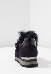 Navy suede sneaker with a glossy black heel accent, textured fur trim, and decorative hardware. Features a grey rubber sole and laces.