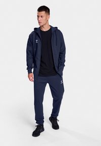 Navy sportswear set includes a hooded jacket and matching pants, made of smooth fabric with a logo detail. Black t-shirt underneath.