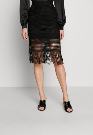 Frau trägt einen schwarzen, strukturierten knielangen Rock mit Fransenabschluss und offene schwarze Mules mit Absatz, stehend vor einem einfarbigen Hintergrund.