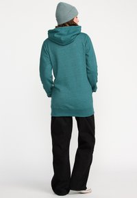 Femme de dos portant un sweat à capuche teal, un bonnet en tricot gris, un pantalon large noir et des baskets blanches, sur fond uni.