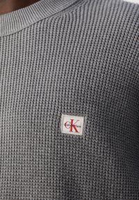 Nahaufnahme eines grauen Strickpullovers mit einem kleinen weißen Patches-Logo, das roten und schwarzen Text "Calvin Klein" auf der Brust zeigt.
