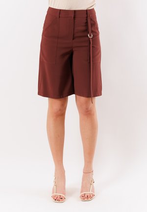 Pantaloncini marrone scuro a vita alta fino al ginocchio con tasche applicate frontali e cintura laterale da annodare, indossati con tacchi aperti beige chiaro.