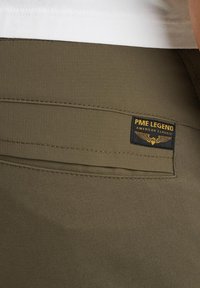 Un pantalon vert olive présente un tissu texturé, des détails de poches cousues et une étiquette PME Legend noire et dorée à l'arrière.