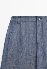 Close-up van blauwe chambray broek met een knoop aan de voorkant, een riemlus en een schuin zakje op een witte achtergrond.