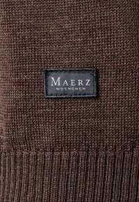 Tessuto di maglione lavorato a maglia marrone con orlo a coste e un'etichetta nera con testo bianco "Maerz Muenchen", che mostra una texture liscia e una tessitura stretta.