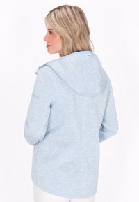 Ljusblå fleecejacka med huva, mjuk textur och en subtil melerad design. Har en logotags på ryggen nära nacken.
