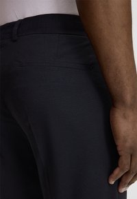 Dunkelblaue Hosen aus einem strukturierten Stoff, mit schmalem Bund und einer Gesäßtasche. Nahaufnahme der Rückennaht und Handplatzierung.