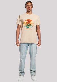 Beige cotton t-shirt featuring a circular graphic in orange, green, and blue depicting mountains and trees. Worn with light blue jeans and white sneakers.