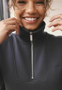 Tmavě šedá mikina s vysokým límcem, předním zipem, strukturální tkaninou a jemným výšivkou značky na hrudi.