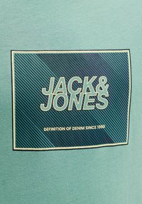 Turquoise t-shirt met een afbeelding met "JACK & JONES" in vet wit op een blauw gestreepte achtergrond, omlijst in lichtgeel.