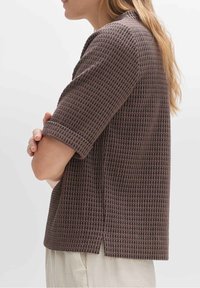 Top corto a maniche corte marrone, con una texture e un sottile motivo a quadri, dotato di spacchi ai lati. Il tessuto sembra leggero e traspirante.