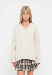 Maglione a V color beige chiaro con maniche oversize, texture morbida e orlo a coste, indossato sopra una gonna a quadri in tonalità di marrone e bianco.