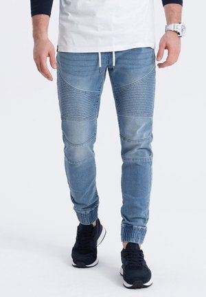 Mann trägt hellblaue gerippte Jogger-Jeans, weiße Kordel, schwarze Sneakers, weiße Uhr und weißes Langarmshirt mit schwarzen Ärmeln.