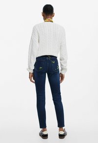 Valkoinen kaapelikudottu neule, tummansiniset farkut, joissa on kukkakirjontaa takataskuissa, sekä kuviolliset tennarit. Malli on selkä menosuuntaan, kädet rentoina.