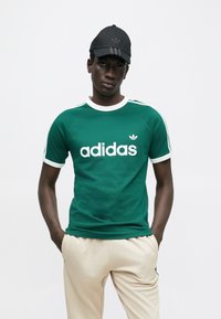 Πράσινο κοντομάνικο μπλουζάκι με λευκό λογότυπο Adidas, γιακά και μανίκια σε αντίθετο χρώμα, συνδυασμένο με μπεζ παντελόνι. Μαύρο καπέλο με τρεις ρίγες.