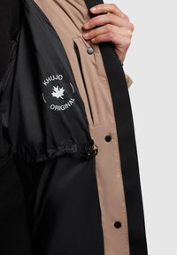 Musta ja beige takki, jonka sisäpuolella on näkyvissä logo "KHUJO ORIGINAL." Varustettu vetoketjullisella taskulla ja kiristysnauhoilla, joissa on klipsit. Sujuva, nylonpintainen.
