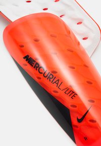 Nike Mercurial Lite sääre kaitsed eredas oranžis, millel on kuusnurksed tekstuurid, must logo ja kõver kuju. Omab perforeeritud valget tagapinda.