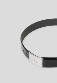 Μαύρη δερμάτινη ζώνη με ορθογώνια ασημένια αγκράφα με ανάγλυφο λογότυπο και λεπτομέρειες ραψίματος. Πέντε ρυθμιζόμενες τρύπες για εφαρμογή.