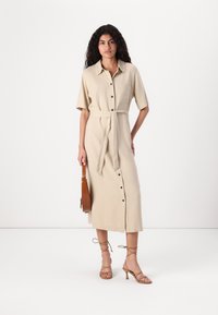 Abito beige con bottoni e maniche corte, cintura a nodo in vita e spacco frontale. Abbinato a sandali con cinturino e una borsa marrone.