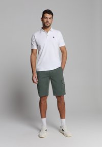 Witte polo met een klein logo, gecombineerd met groene cargoshorts met zijzakken. Model draagt witte sneakers met blauwe accenten.