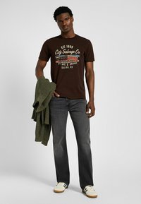 Camiseta de algodón marrón oscuro con gráfico frontal, combinada con jeans grises y zapatillas blancas con rayas negras. Suéter de punto verde colgado del brazo.
