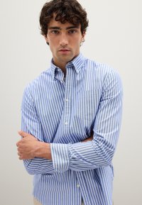 Camisa de botones a rayas azul y blanca con cuello, mangas largas y un bolsillo en el pecho; hecha de una tela ligera con una textura suave.