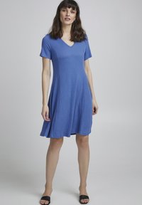 Kurzes, kurzärmliges blaues Kleid aus strukturiertem Stoff mit V-Ausschnitt und ausgestelltem Schnitt, das oberhalb des Knies endet. Getragen mit schwarzen Sandalen.