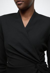 Čierne zavinovacie šaty z hladkej, textúrovanej látky s hlbokým výstrihom do V a prekrúteným detailom v páse.