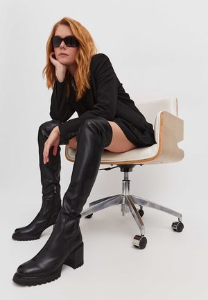 Black thigh-high leather boots with a chunky heel and textured soles. Model wears a black blazer and sits in an ergonomic chair.