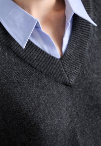 Maglione grigio in lana con design a scollo a V sovrapposto a una camicia col colletto azzurro chiaro. Texture a coste visibile lungo il colletto e l'orlo.