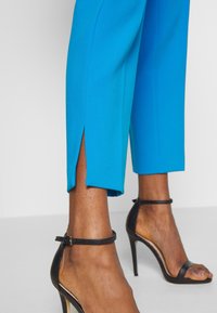 Dolne nogi ubrane w jasnoniebieskie ścięte spodnie z małymi rozcięciami i czarne sandały na wysokim obcasie z paskami na kostce na białej podłodze.