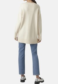 Pull oversize en tricot crème avec des épaules tombantes, un ourlet côtelé et des manches longues, associé à un jean droit bleu et des chaussures argentées.