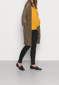 Těhotná žena v žlutém topu, hnědém pleteném kardiganu, černých skinny džínách a černých mokasínech stojící na pozadí jednoduché bílé stěny.