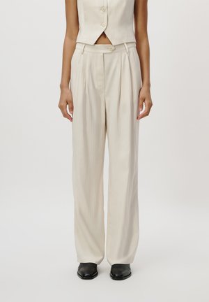 Beige ermeløs vest med rund hals, kombinert med høy midje med plisserte bukser. Begge plaggene har en glatt, lett tekstur.