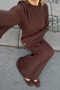 Robe en tricot marron texturée avec des manches longues, un col rond et une coupe ample, portée avec des chaussures plates marron sur une surface en pierre.