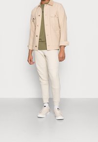 Beige canvasjas met borstzakken, olijfgroene t-shirt, crème joggingbroek en lichte schoenen met gestructureerde veters. Geen zichtbare patronen.