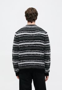Person with short curly hair seen from behind wearing a black and gray striped fuzzy sweater and black pants against a white background.