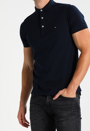 Mann trägt ein marineblaues, kurzärmeliges Poloshirt mit kleinem Logo auf der Brust und schwarze Jeans, rechte Hand in der Tasche, vor weißem Hintergrund.