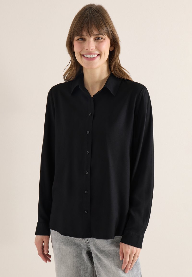 Femme souriante portant une chemise noire à manches longues boutonnée et un jean gris clair, debout devant un fond beige uni.