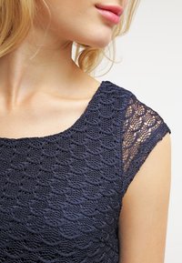 Robe en dentelle bleu marine avec un motif festonné et des manches courtes, mettant en valeur des détails texturés complexes le long de l'épaule.