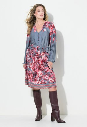 Blumenkleid in Grau mit pinken Blumen, langen Ärmeln, V-Ausschnitt, Knopfdetails, Taillengürtel und knielanger Rock. Kombiniert mit braunen knielangen Stiefeln.