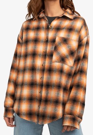 Vrouw die een oversized oranje, zwarte en witte geruite blouse met borstzak en een blauwe spijkerbroek draagt, staand tegen een witte achtergrond.