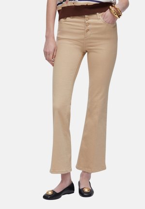 Pantaloni beige a vita alta con fondo svasato e quattro bottoni, indossati da una persona con ballerine nere decorate con cerchi dorati.