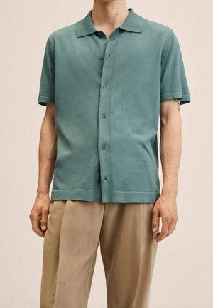 Mężczyzna w krótkim, zapinanym na guziki turkusowym koszuli i beżowych spodniach, stojący na tle jednolitego jasnego tła.